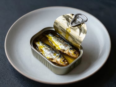 Top-down editorial shot of an open gourmet tin of sardines in golden olive oil, placed on a minimalist ceramic plate in a Scandinavian dining setting.