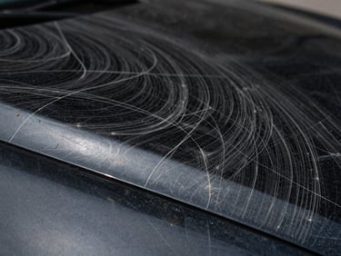 Macro extreme close-up of a scratched, swirly black car paint under harsh white LED spotlight, showing heavy spiderweb scratches and oxidation.