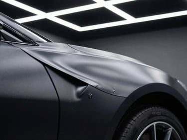 Macro shot of a self-healing polyurethane film being stretched over a matte black hypercar fender, high-contrast studio lighting, bright white LED overhead grid lines reflecting perfectly on the surface.