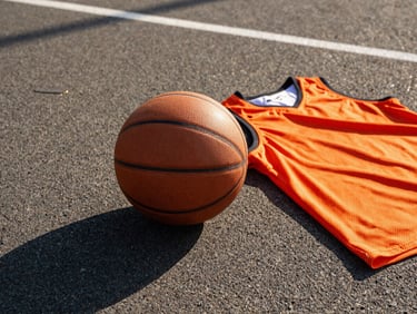 Close-up of a brand new basketball and orange mesh jerseys resting on an asphalt court, harsh midday sun.