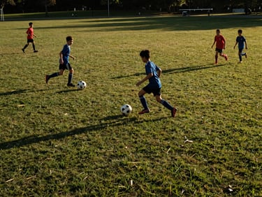 Wide-angle action shot of kids playing soccer on a public park grass field, natural golden hour lighting.