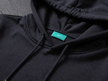 Moody low-key studio shot of a dark slate folded hoodie with a custom woven electric teal neck tag stitched cleanly into the collar.
