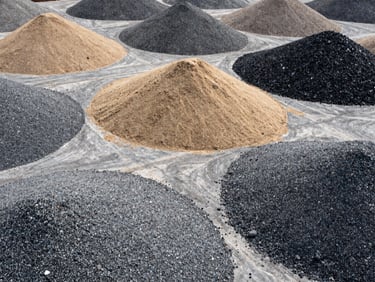 Overhead flat-lay of construction aggregate materials — crushed stone, sand, gravel piles on a warehouse floor, natural overcast daylight, high-contrast texture, no people