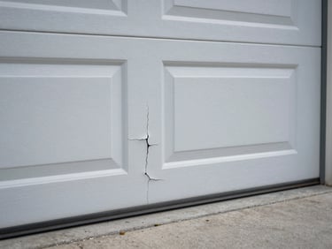 Close-up of a bent garage door panel section showing a deep crease and paint cracking along the fold, natural outdoor light, concrete driveway visible below