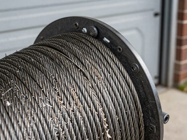 Close-up of a frayed steel cable looped around a drum at the base of a garage door, harsh natural light revealing individual wire strands beginning to separate