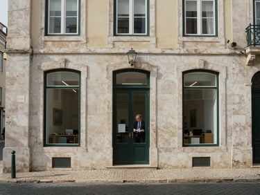 Wide shot of a Lisbon stone-facade building housing a legal office, viewed from across a quiet cobblestone street in flat overcast morning light, a professional visible through a ground-floor window reviewing papers