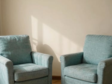 A quiet, sunlit therapy room with two comfortable linen armchairs facing each other, soft leaf shadows on a warm cream wall