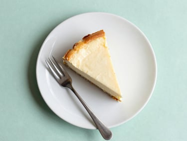 Overhead flat-lay of a single slice of creamy cheesecake with a golden crust on a matte white ceramic plate, a delicate silver fork beside it, soft natural daylight, mint green background, 35mm