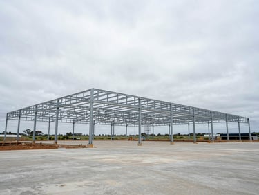 Wide-angle ground-level view of a commercial agricultural facility under construction on the KwaZulu-Natal south coast, steel portal frame rising against an overcast sky, concrete slab visible, no workers posed