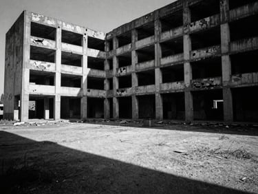 Wide, isolating black and white environmental shot of decaying brutalist architecture under harsh sunlight, long shadows, surrealist composition.