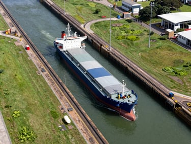 Overhead view of the Panama Canal locks from the air, a cargo vessel mid-transit, green banks on both sides, clear daylight, wide environmental frame showing the operational scale of the waterway