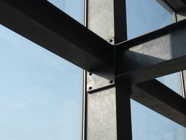 Ultra-tight crop of a structural concrete joint and blackened steel beam intersecting a massive glass pane, natural high-contrast morning light.