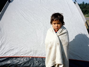 A young child wrapped in a warm blanket standing next to an emergency shelter tent, authentic photojournalism