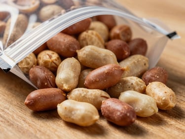 Macro close-up of mixed flavored peanuts in a clear sealed bag on a natural wood surface, warm directional light showing coated textures and color variety