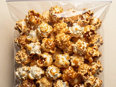 Close-up overhead flat-lay of gourmet caramel popcorn kernels in a sealed transparent bag on a warm-white surface, warm studio light highlighting caramel gloss and texture