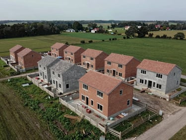 Panoramic drone aerial of a mid-construction housing development, multiple house plots at different build stages visible, concrete foundations and brick courses rising, surrounding green fields, overcast daylight, no people, muted earth and grey tones