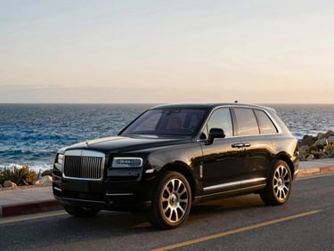 Wide cinematic shot of a clean, black luxury SUV parked on a quiet coastal road in Los Cabos during golden hour, the deep blue Pacific ocean in the background, soft warm light reflecting off the polished vehicle.