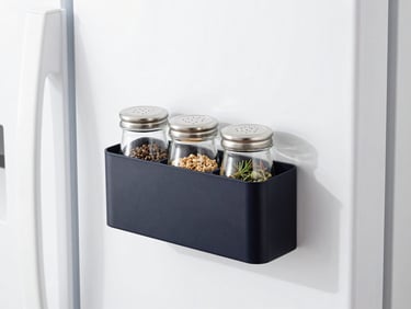Close-up of a black magnetic kitchen organizer holding glass spice jars on a white refrigerator side, bright daylight, minimalist kitchen setting.