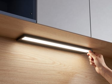 Hands installing a slim magnetic LED light bar under a modern wooden cabinet, soft warm glow illuminating the counter, clean minimalist aesthetic.