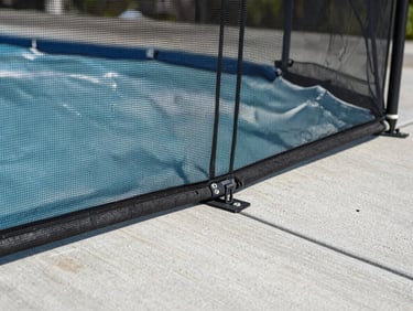 Close-up environmental shot, a section of removable mesh pool fence anchored into a concrete deck, natural daylight from above, camera at child's eye level showing the fine mesh weave and the tamper-resistant sleeve anchor flush with the deck surface