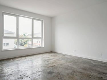 Wide interior shot of a bright apartment living room, white walls, large window with natural daylight, polished concrete floor, no furniture staging, clean and honest condition