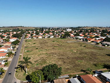 Open land plot in a Brazilian suburban neighborhood, aerial perspective at low angle, visible street access, clear sky, surrounding residential context