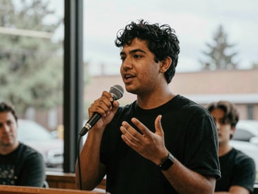 Candid medium shot of a young Seattle advocate speaking passionately into a microphone at a neighborhood forum, natural overcast daylight filtering through window, expressive hand gestures