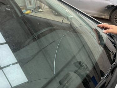 Close-up of a technician's gloved hands pressing a repair resin tool into a windshield crack under bright side-raking overhead light, glass surface sharp and clear
