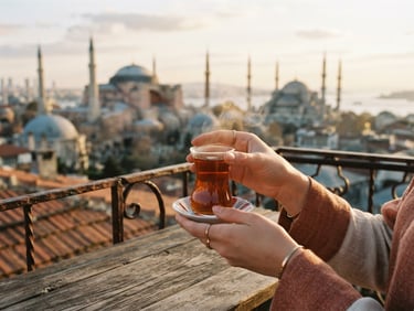 Close-up of elegant hands holding a traditional Turkish tea glass, warm steam rising, soft morning light, blurred background of historic Istanbul minarets, warm terracotta tones, 35mm film look