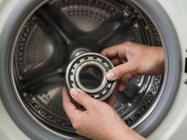 Technician hands removing a washing machine drum bearing in a home laundry room, workshop lamp casting directional light on the steel drum and worn bearing ring
