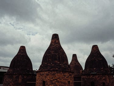 Atmospheric, high-contrast 35mm photograph of brick kilns in the distance under a heavy, cloudy sky, desaturated tones, deep shadows.
