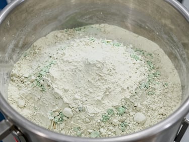Overhead view of a pharmaceutical powder blending vessel interior, fine white-and-green powder blend uniformly distributed, stainless steel walls, bright facility lighting, no people