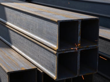 Thick structural steel beams stacked in a fabrication yard, daylight reflecting off metallic edges.