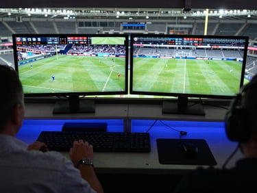 Overhead production-deck shot of a league scoreboard operator at a broadcast desk, dual monitors showing live match graphics, stadium tier visible through glass above, arena lighting casting blue on equipment panels