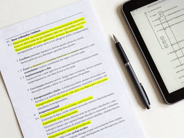 Overhead flatlay of an organized desk with a printed Portuguese exam sheet, yellow highlighter marking key terms, a sleek pen, and a tablet displaying a logical syntax diagram, clean natural daylight