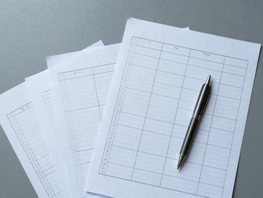 Overhead flat-lay close-up of organized planning sheets, a ruled checklist, and a slim pen arranged on a cool-grey surface, cool north-facing daylight, no hands visible, precise and orderly arrangement, generous empty space in the upper left
