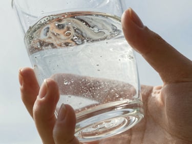 Extreme close-up under bright overexposed midday sun showing cloudy, textured liquid in a glass, held by hands with chipped nail polish.