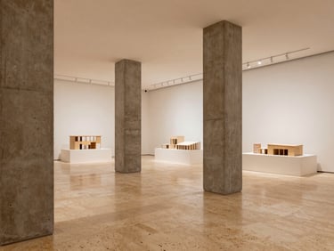 Architectural presentation gallery, warm travertine floors, raw concrete columns, physical architectural models under soft diffused daylight, minimal layout