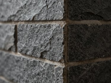 Close-up of cut stone tile edge at a wall corner, mortar bed visible, raking side light showing surface texture and material density, dark grey tones