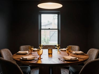 Wide environmental shot of a private dining room setup — intimate round table for eight, dark walls, single pendant light casting a warm pool, hand-folded napkins, amber glassware, no guests, afternoon light through a narrow window