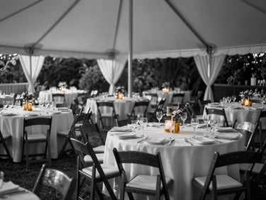 Panoramic view of an outdoor wedding reception setup, round tables with white linen and gold candleholders under a marquee, late-afternoon directional daylight, empty chairs awaiting guests, black-and-white with single warm amber candle glow
