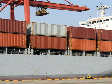 A commercial cargo ship docked at a port, steel containers being loaded under bright daylight.