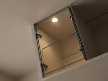 Overhead 45-degree view of a built-in glass-fronted storage unit recessed into a residential wall, sharp focus on the frameless glass door hinge and interior shelf edge, warm overhead light, muted neutral wall finish
