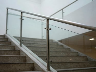 Wide-angle view of a frameless structural glass balustrade at the edge of an interior staircase, sharp focus on the stainless point-fix fitting at mid-span, cool diffuse daylight from above, warm concrete stair treads visible below