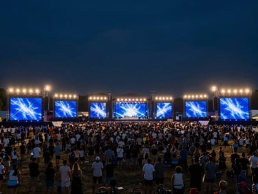 Wide panoramic shot of a completed outdoor festival stage at night, full production rig lit in deep blue and white stage wash, crowd of tens of thousands filling the field, practical floodlights on the horizon