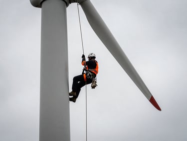 Technician in full safety gear, rope-access certified, rappelling down a wind turbine tower, dramatic low-angle shot against an overcast sky, high-contrast desaturated tones with safety orange accents.