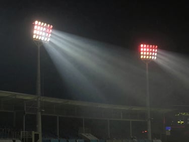 Dynamic cricket stadium floodlights cutting through mist, dark cinematic atmosphere, crimson accents, 35mm.