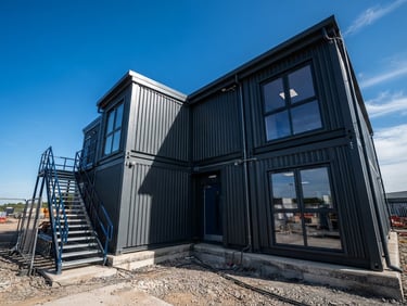 Low-angle architectural photograph of a high-spec modular site office clad in charcoal grey corrugated steel, bright daylight casting sharp geometric shadows.