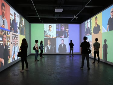 Wide shot of a darkened immersive research installation room, participants standing inside surrounded by projected consumer footage and emotion-coded color overlays on walls, candid documentary framing, low ambient light