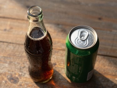 Overhead close-up of a glass soda bottle and a can on a worn wooden surface, golden afternoon light from the left, condensation visible on the bottle